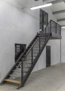 Ocelové schodiště se širšími nášlapy a zábradlím, umístěné v industriálním prostoru.